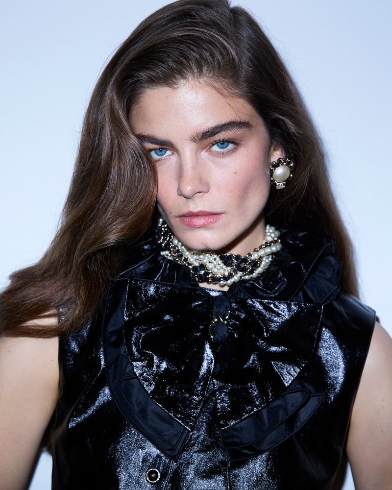 Portrait of a stylish woman with brown hair, wearing a black patent leather vest with a ruffled collar and a pearl necklace and earrings.