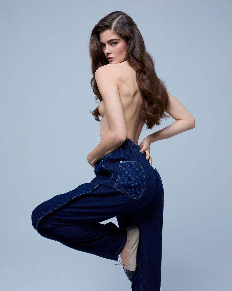 Brunette woman in blue jeans and ballet flats poses in a studio with a confident expression, looking over her shoulder at the camera.