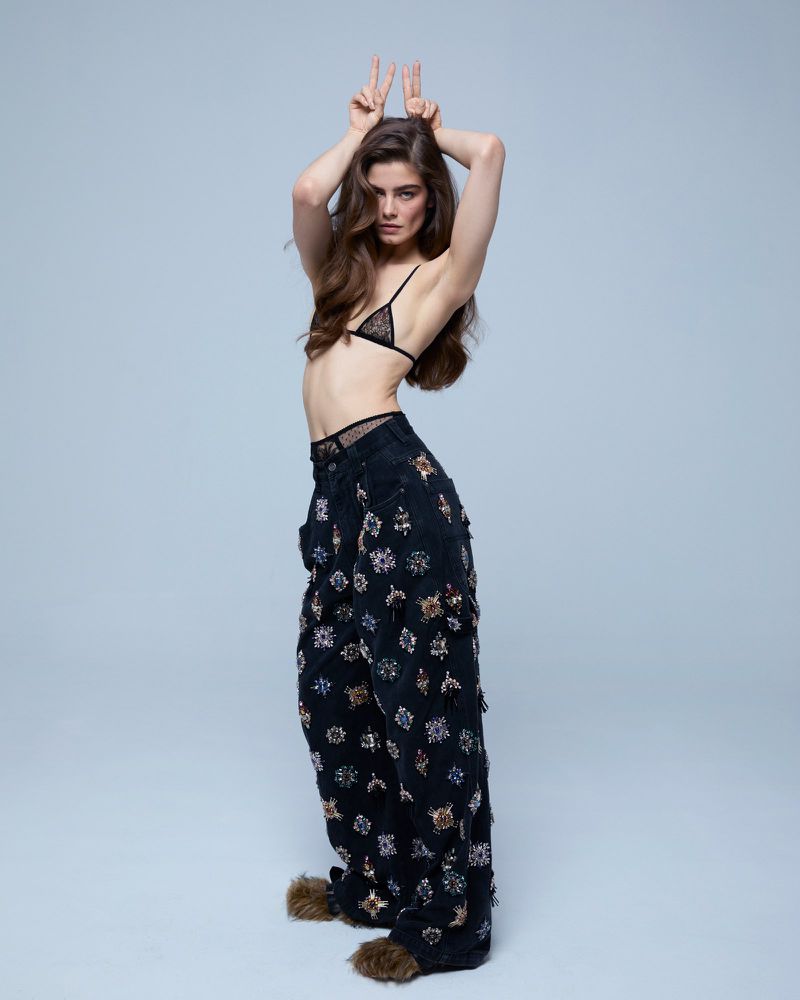 A woman in black bikini top and floral pants poses in a studio with her hands up making peace sign gestures in a full shot.