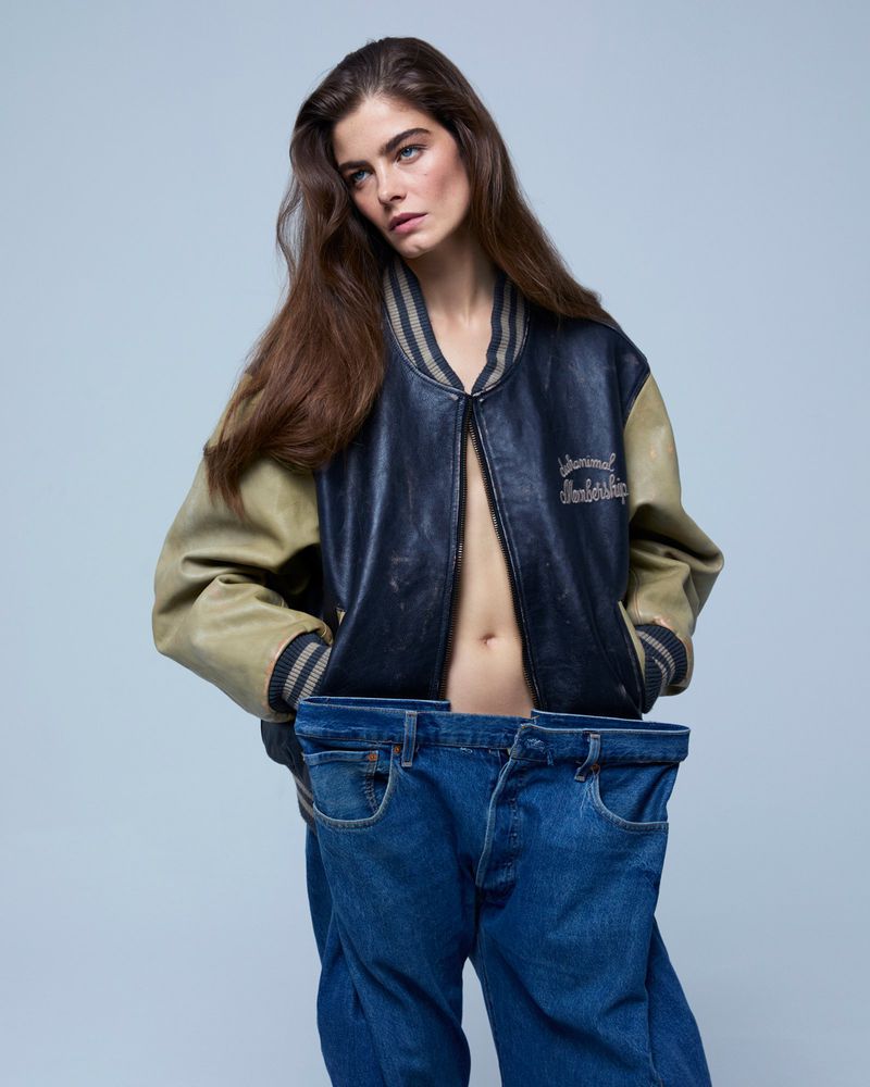 Brunette adult woman wearing a vintage varsity jacket, cropped to show denim jeans being held up in front of her, studio shot.