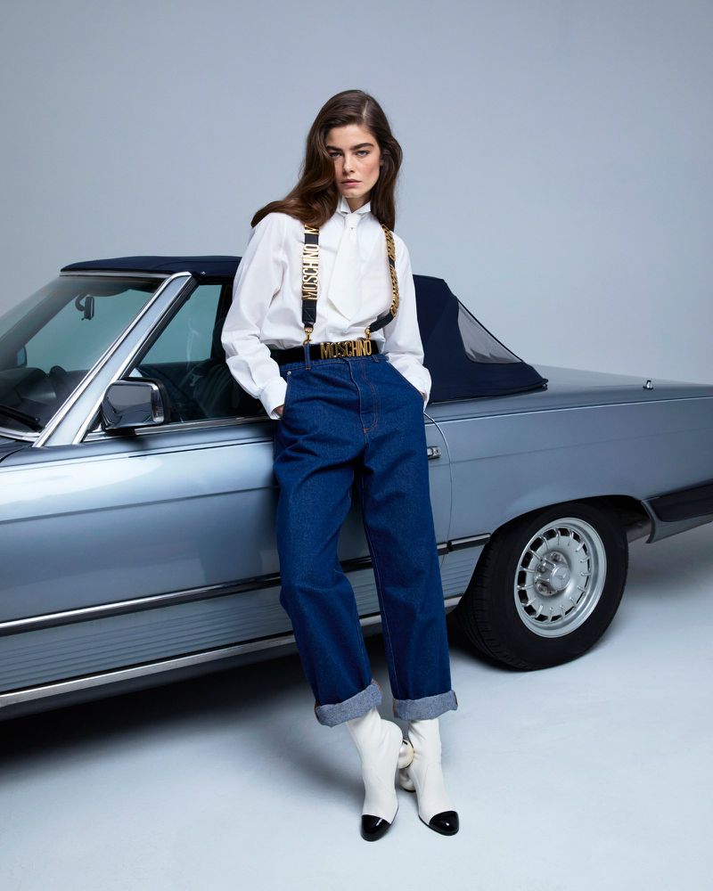 Fashion shot of a woman leaning against a light blue convertible car in a studio, wearing a white shirt, jeans, and white boots.