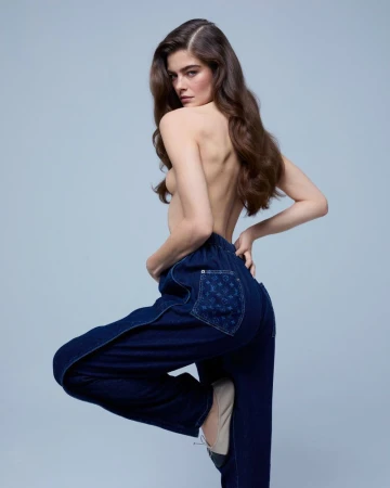 Model Merlijne Schorren posing topless from behind in dark blue denim trousers against a light blue-grey studio backdrop.