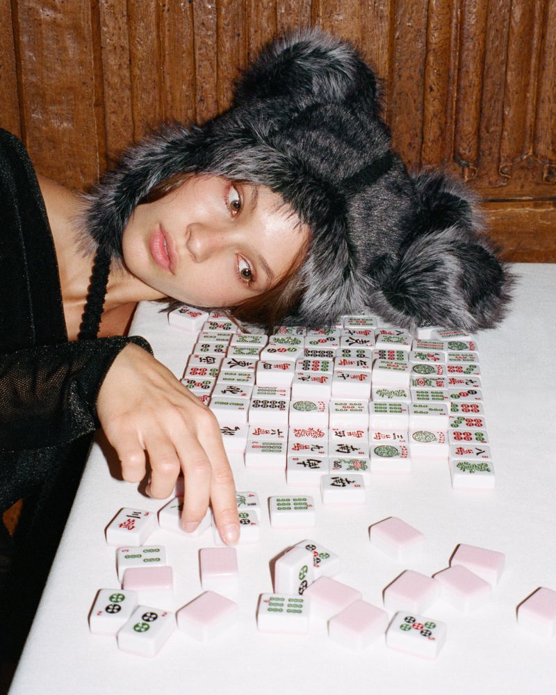 Young woman in fur hat plays mahjong on white table, indoor setting, warm light, black top, focus on the game and her expression.