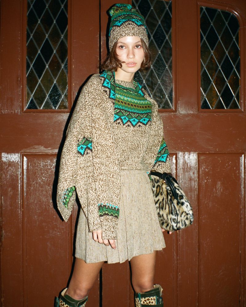 Woman in a patterned knit sweater and matching hat stands in front of a dark wooden door with a patterned skirt and animal-print boots.
