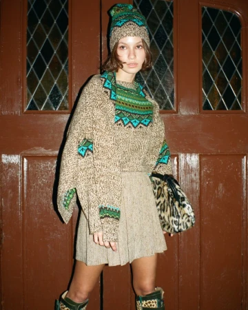 Woman in a patterned knit sweater and matching hat stands in front of a dark wooden door with a patterned skirt and animal-print boots.