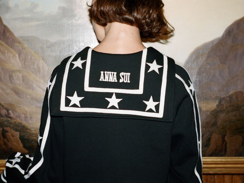 A person with short brown hair wears a black top with white star details and an Anna Sui logo against a blurred painting backdrop.