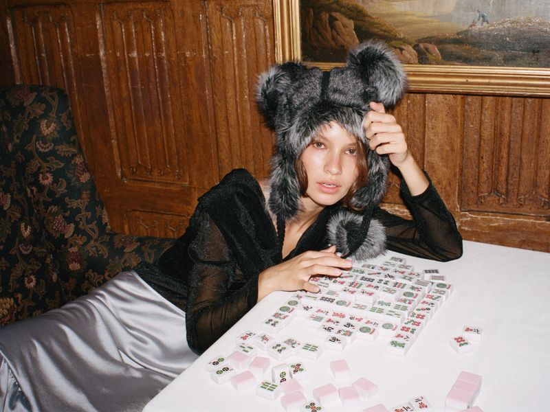 Stylish woman in faux-fur hat poses at a table with Mahjong tiles, creating a vintage-style fashion portrait indoors with a rich, warm palette.