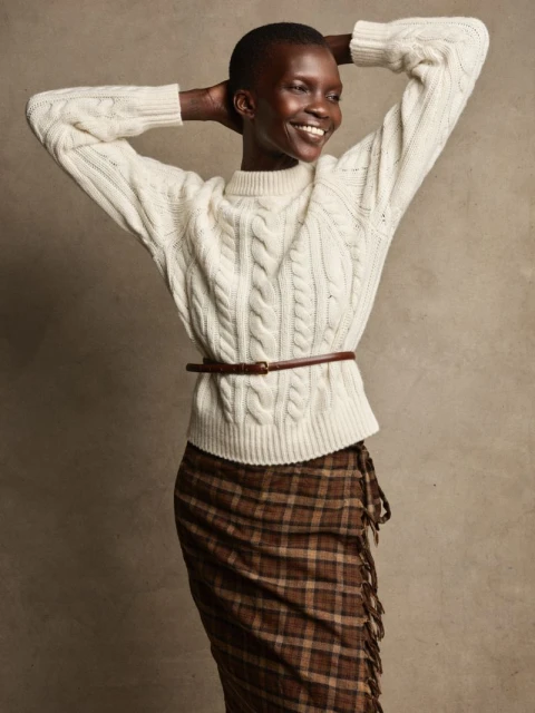 Woman posing with hands behind her head, wearing a cable knit sweater, brown belt, and plaid skirt in a studio setting.