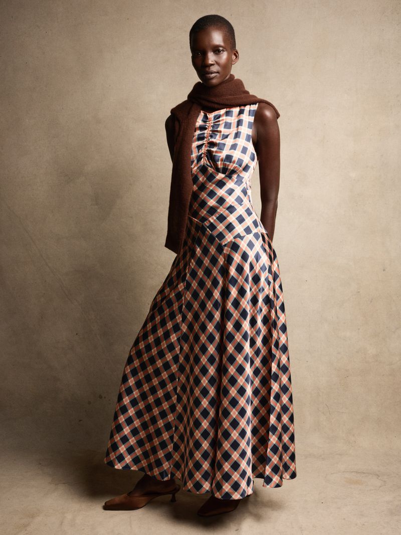 Full-length shot of a Black woman in a sleeveless diamond-pattern dress with a brown scarf, posing against a neutral backdrop.