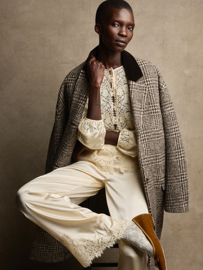 Woman in silk lace-trimmed pajamas and plaid wool coat poses indoors against a neutral backdrop in soft, diffused lighting.