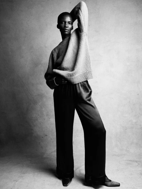 Monochrome fashion portrait of an adult woman in a sweater and trousers posing in a studio with hand above her head.