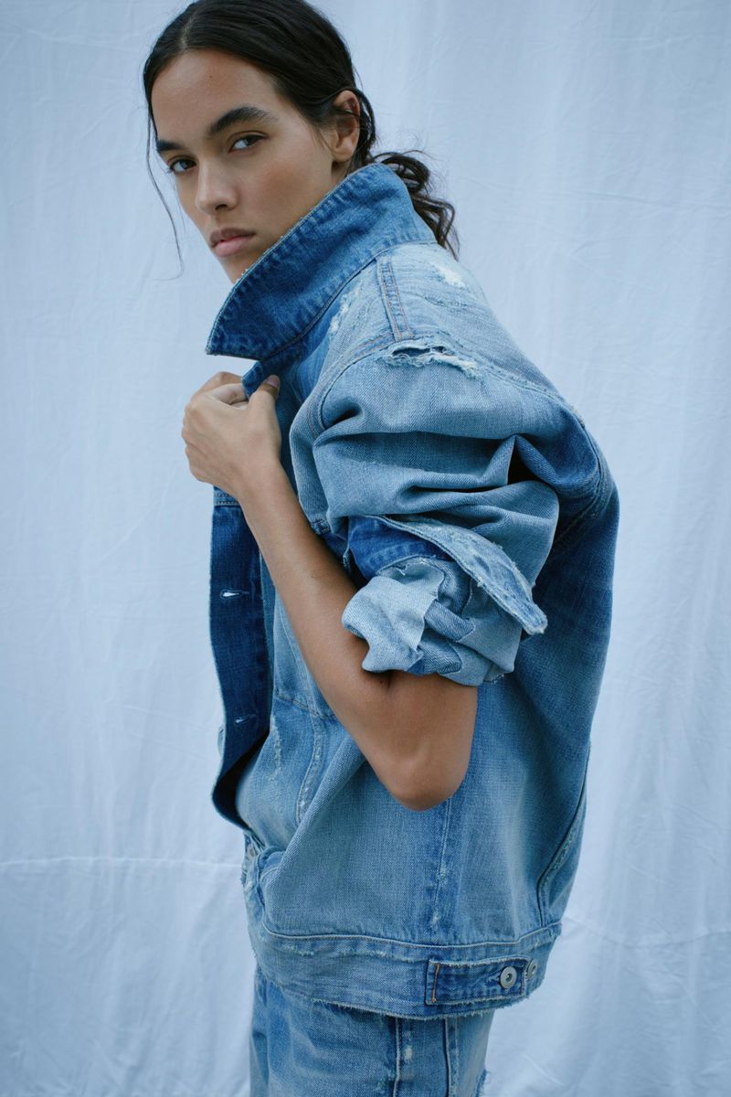 A woman poses in a distressed denim jacket and jeans against a simple light background in a fashion forward portrait.