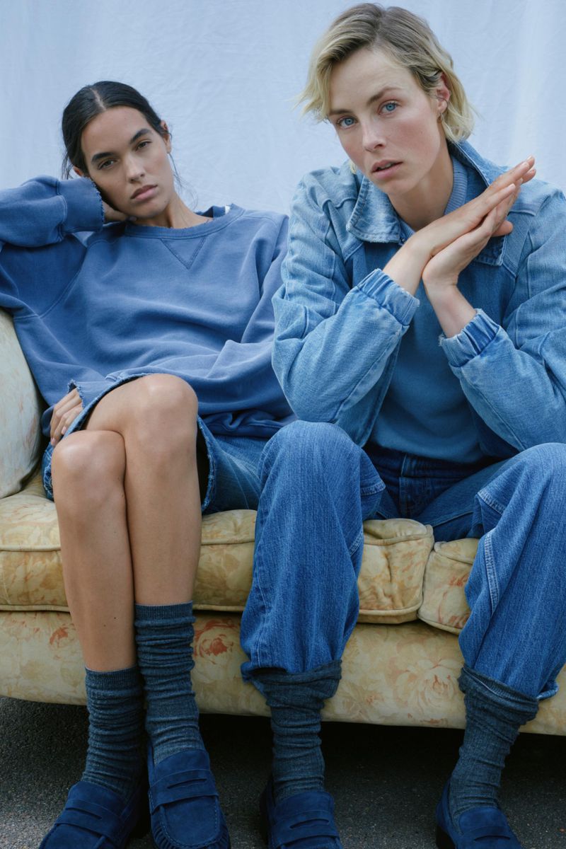 Two women in blue outfits sit together on a couch. One wears a denim jacket and jeans, the other a sweatshirt and shorts.