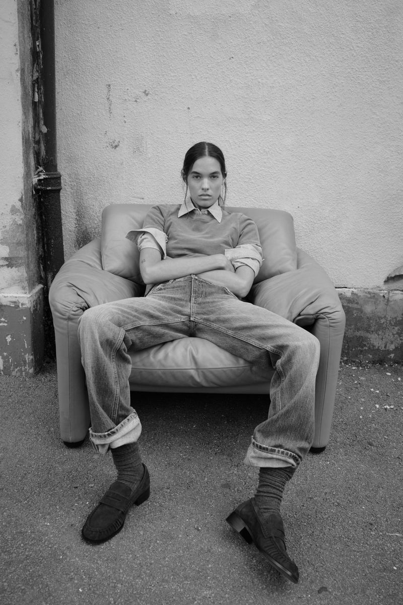 Monochrome shot of a woman in denim jeans and loafers sitting with arms crossed in an armchair outdoors against a textured wall.