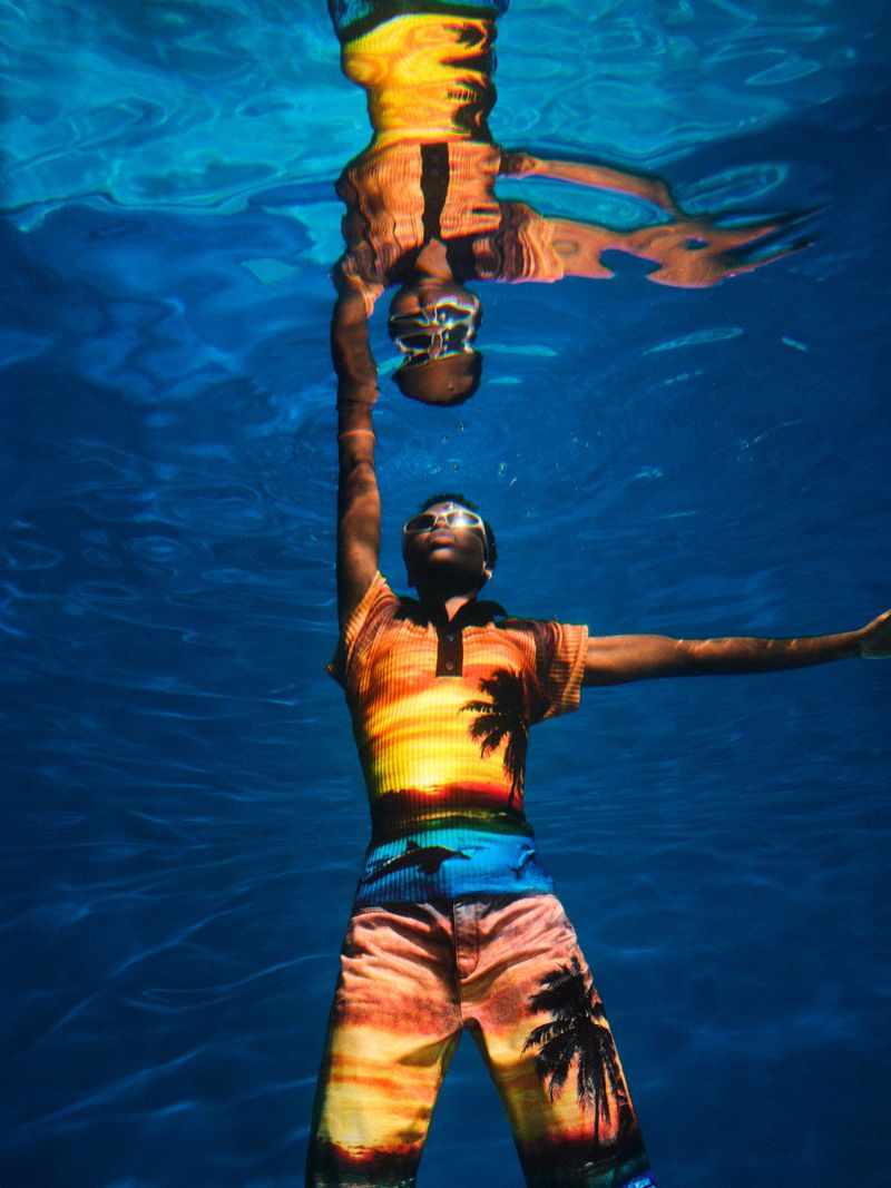Underwater shot of a man in patterned resort wear with arms outstretched, mirrored in the water's surface with a blue water background.