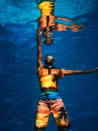Underwater shot of a man in patterned resort wear with arms outstretched, mirrored in the water's surface with a blue water background.
