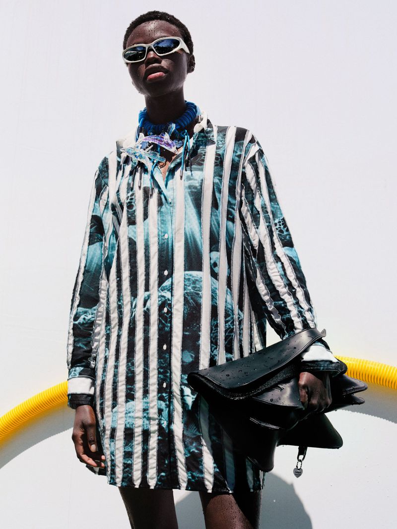 A fashionable African-American woman models a striped shirt-dress with a handbag against a bright backdrop, showcasing fashion and style.