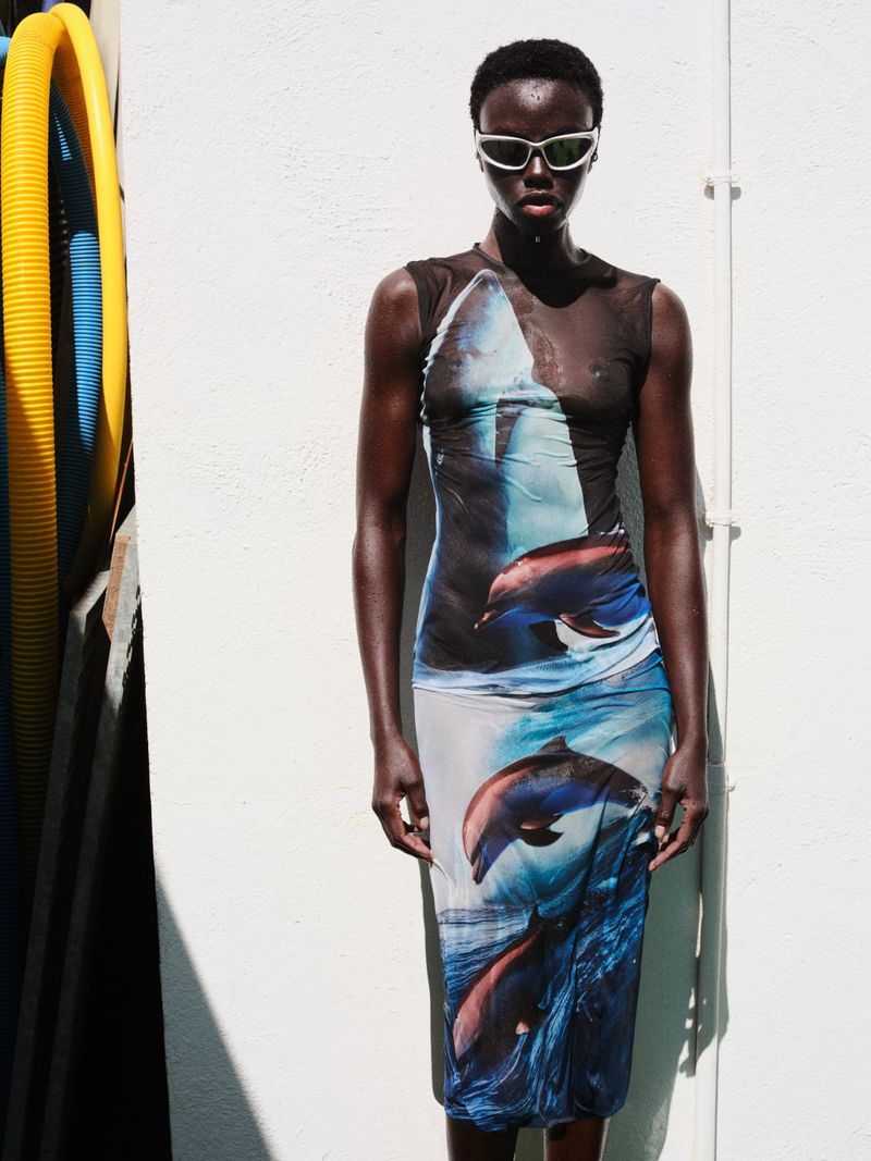 Adult female fashion model wearing a sleeveless midi dress with a dolphin print stands in front of a white wall with blue pipes.