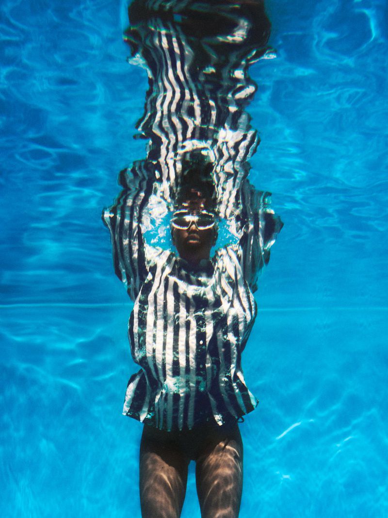 Underwater shot of an adult female wearing dark sunglasses and a striped blouse in a turquoise swimming pool with clear water.
