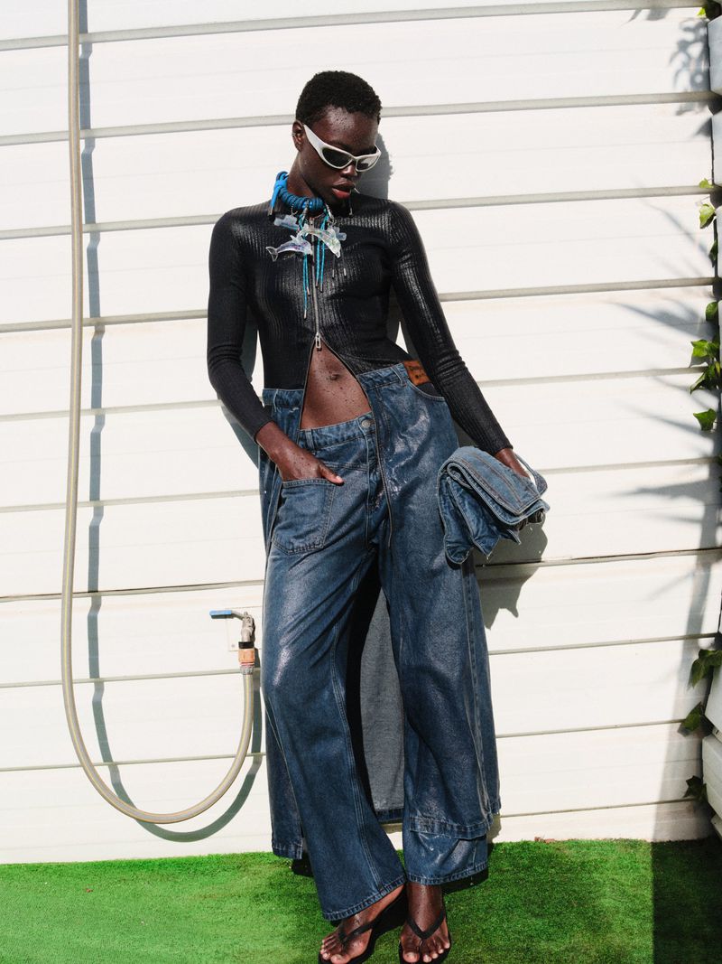Fashion shot of a Black woman in wide-leg denim pants and a black top poses outdoors against a white wall in sunlight.