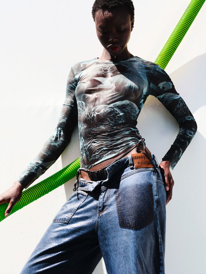 Fashion shot of a Black female adult wearing a patterned long-sleeve top and wide-legged jeans leaning against a white wall with a green tube.