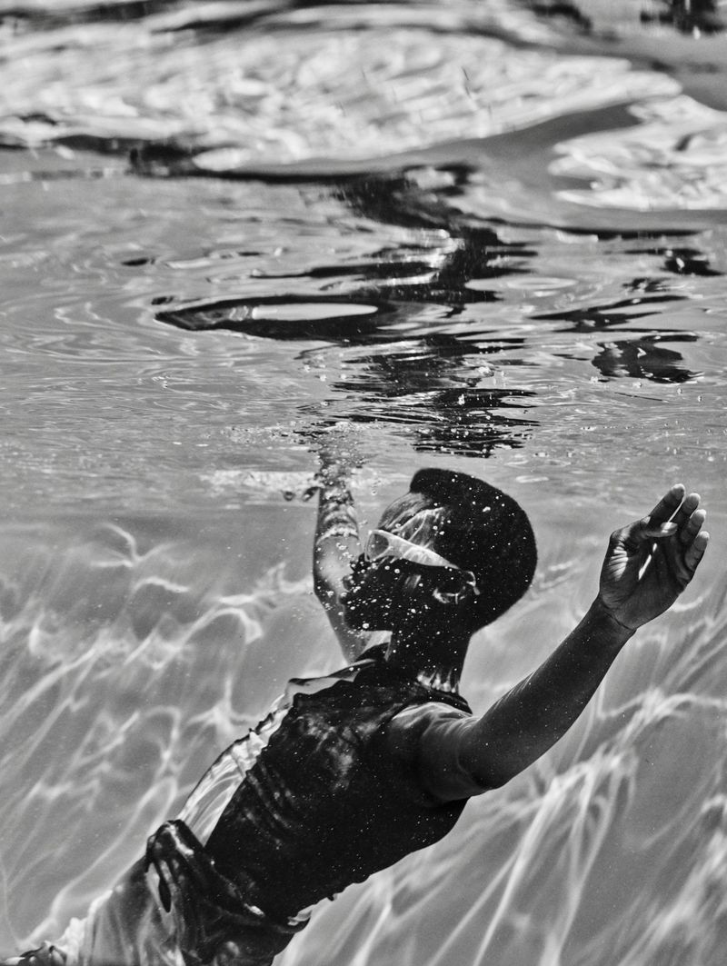 Underwater view of a child swimming, monochrome tones, showing arms outstretched, rippled water surface, and natural light.