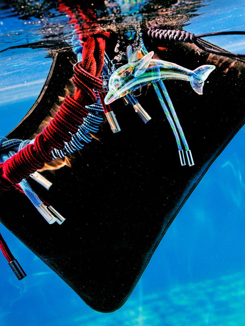 A black purse with colorful rope details and a dolphin accessory is submerged in bright blue water creating a surreal, underwater fashion statement.