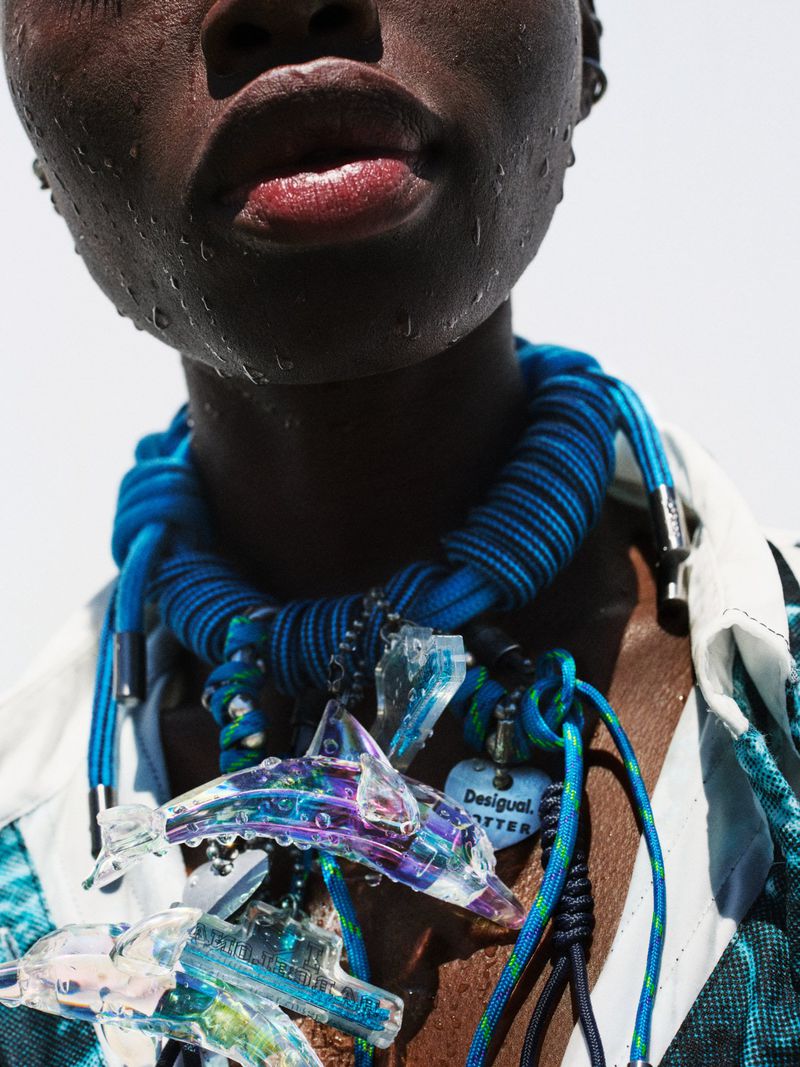 Close-up of a dark-skinned adult woman wearing a blue rope necklace with clear dolphin pendants and a patterned shirt against a white background.