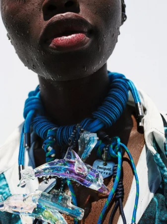 Close-up of a dark-skinned adult woman wearing a blue rope necklace with clear dolphin pendants and a patterned shirt against a white background.