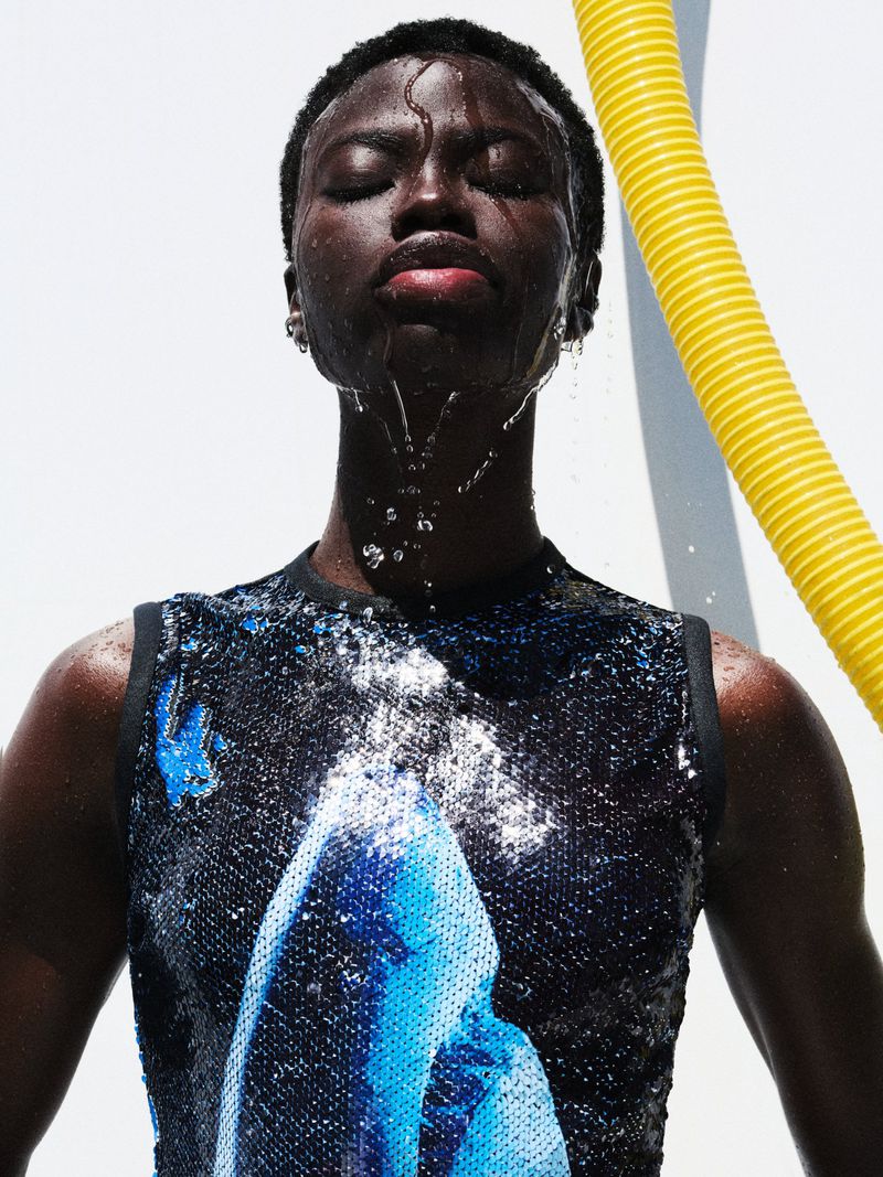 Portrait of Black male model with water cascading over his face, wearing sequined top, set against a bright white background with yellow tube.