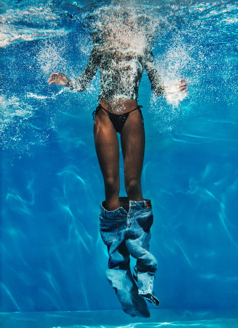 Underwater shot of a woman in a bikini top with jeans partially on her legs, creating an abstract and surreal effect in blue water.