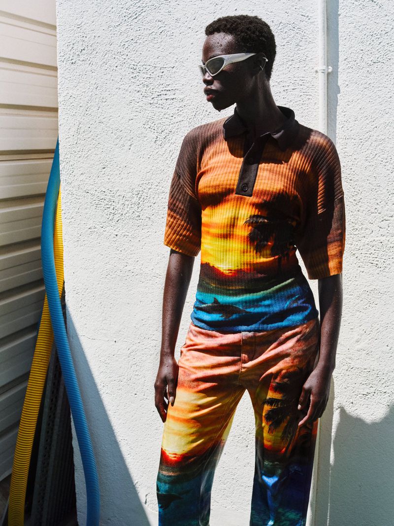 Dark-skinned woman wearing sunglasses and patterned matching top and pants poses against a white wall in bright sunlight outdoors.