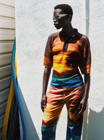 Dark-skinned woman wearing sunglasses and patterned matching top and pants poses against a white wall in bright sunlight outdoors.