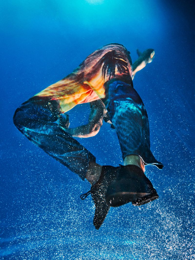 Person in colorful pants underwater, captured from a low angle with visible water droplets and sunlight filtering through the water.