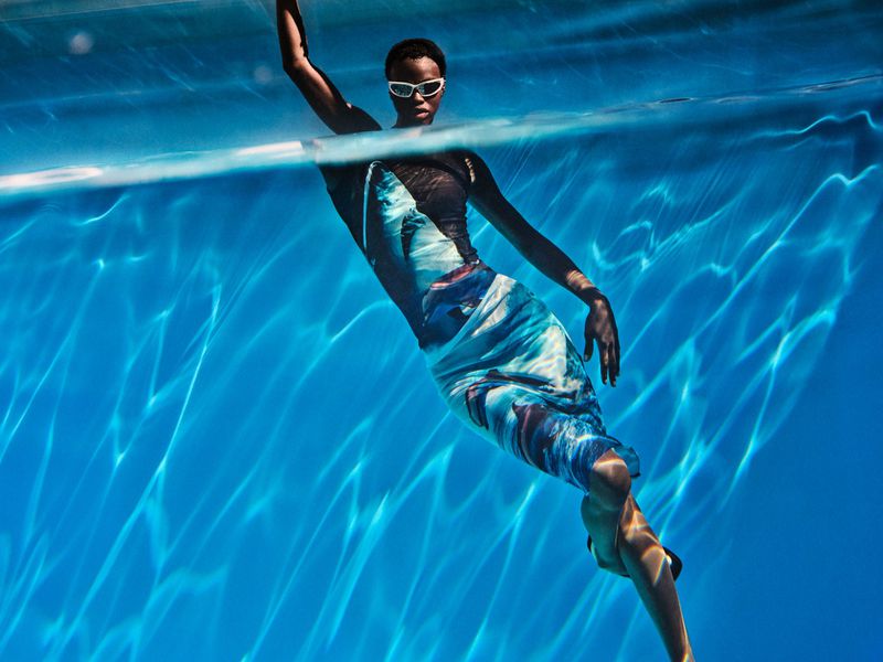 Underwater shot of an African-American woman in a blue dress and white goggles swimming in a pool with sunlit water reflections.