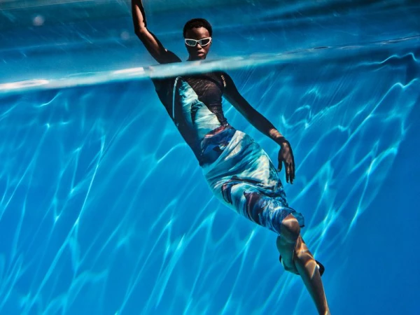 Underwater shot of an African-American woman in a blue dress and white goggles swimming in a pool with sunlit water reflections.