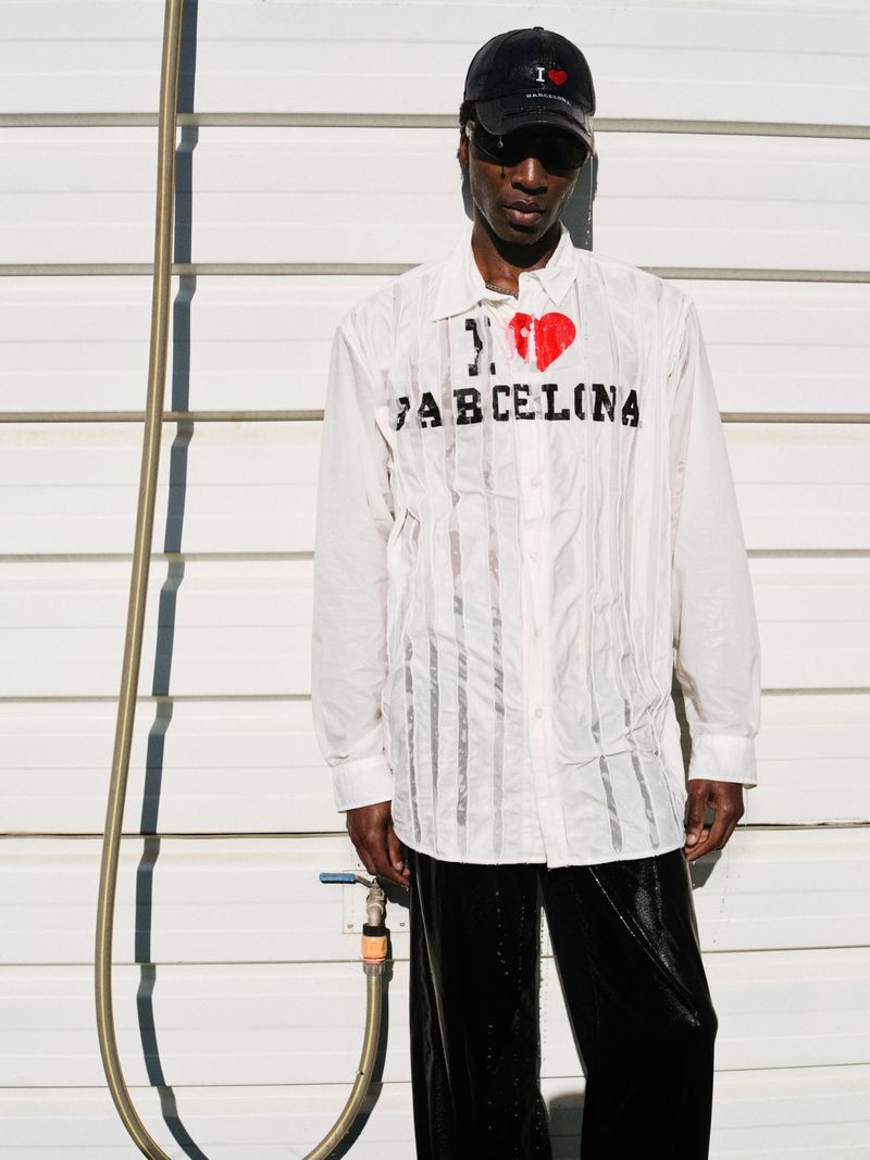 Adult male in sunglasses and a baseball cap stands in front of a white wall wearing an 'I Heart Barcelona' shirt, in an outdoor setting.