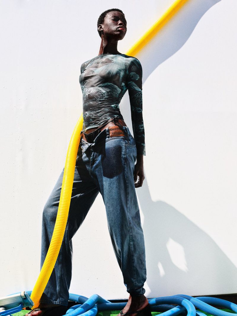 A dark-skinned woman poses outdoors in a printed top and jeans, framed by yellow and blue tubing against a stark white wall in sunlight.