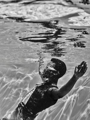 Black-and-white underwater image of a model floating submerged in water with light caustics playing across the surface and pool floor.