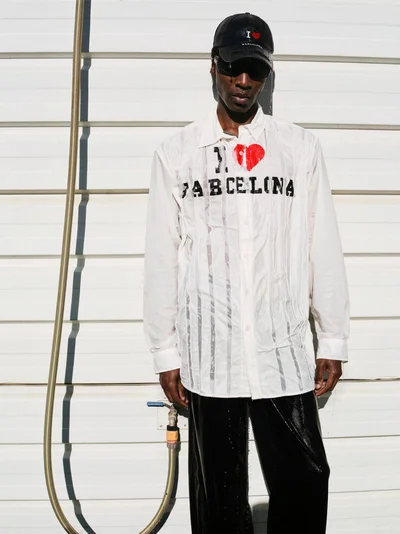 Model wearing white printed shirt and black trousers standing against a white paneled wall with a hanging hose