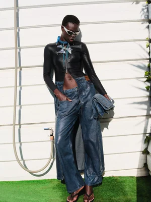 Model standing against a white slatted wall wearing a black zip top, blue denim trousers, and futuristic sunglasses.
