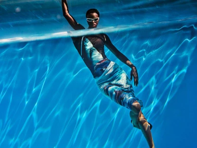 A model wearing white-framed sunglasses and a blue printed dress floating submerged underwater in a sunlit swimming pool.