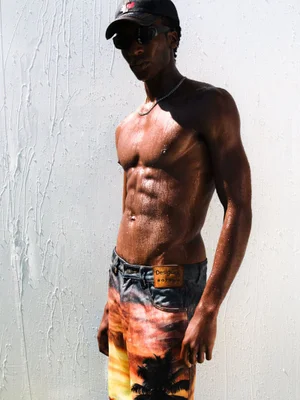 Shirtless male model wearing printed jeans, sunglasses, and a cap against a wet white wall