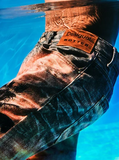 Close-up of person wearing denim jeans submerged in vibrant blue water with a branded leather waist patch.