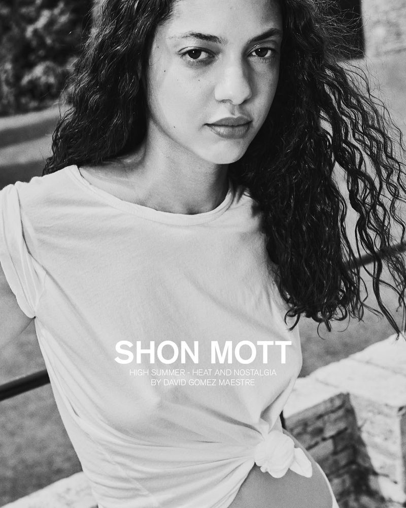 A portrait of a young woman with curly hair in a white t-shirt posing outdoors in a black and white photograph.