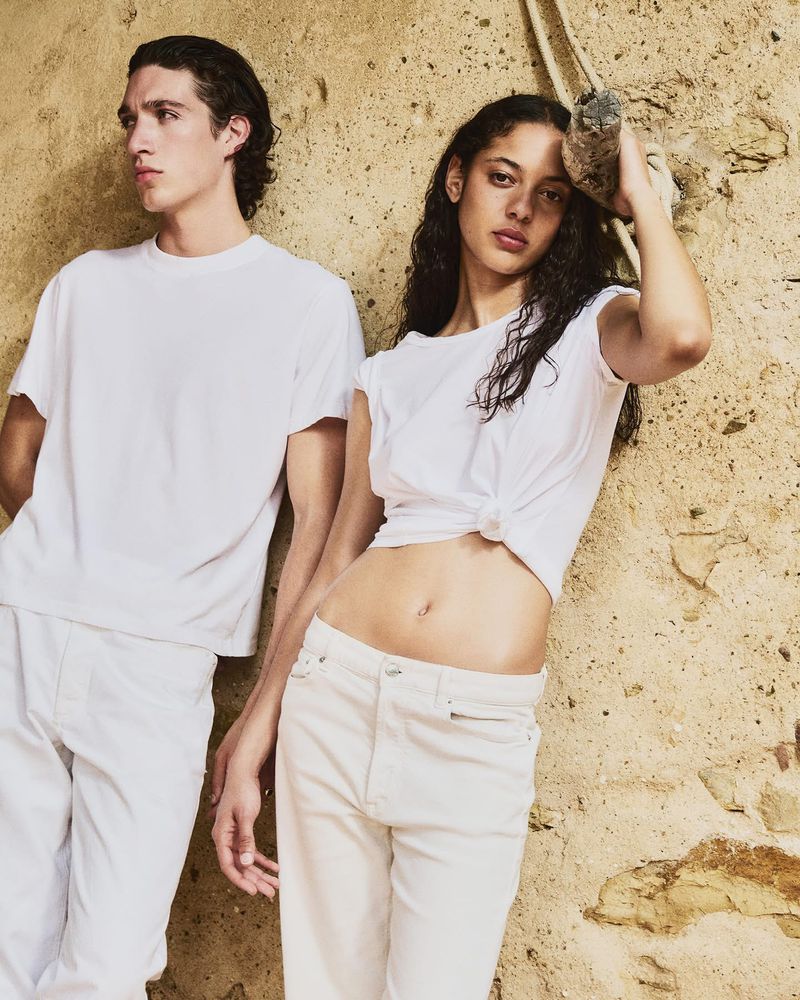 A stylish couple poses against a textured wall wearing white t-shirts and light denim jeans in soft, natural light. Fashion shot, studio.