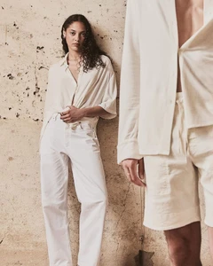 Model Annabelle Pouilly leaning against a textured stone wall in cream shirt and white trousers for Shon Mott.