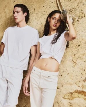 Two models wearing white t-shirts and trousers leaning against a textured stone wall outdoors.