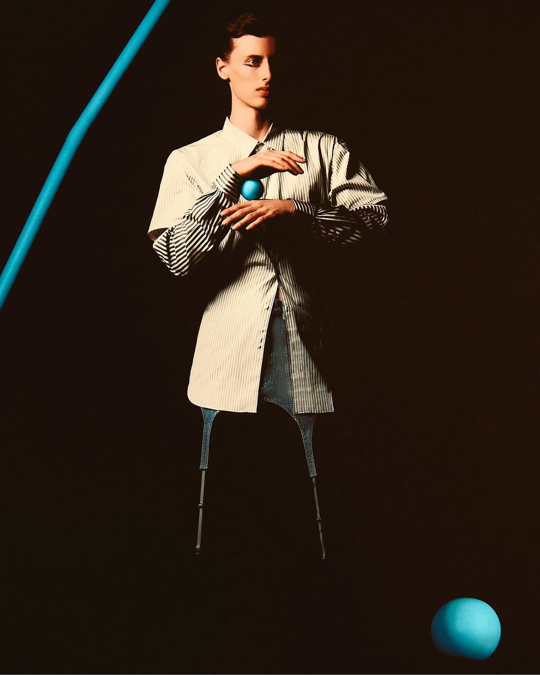 Stylish adult in shirt and blue pants posing with blue sphere against dark backdrop in studio fashion shot with blue pipe prop.