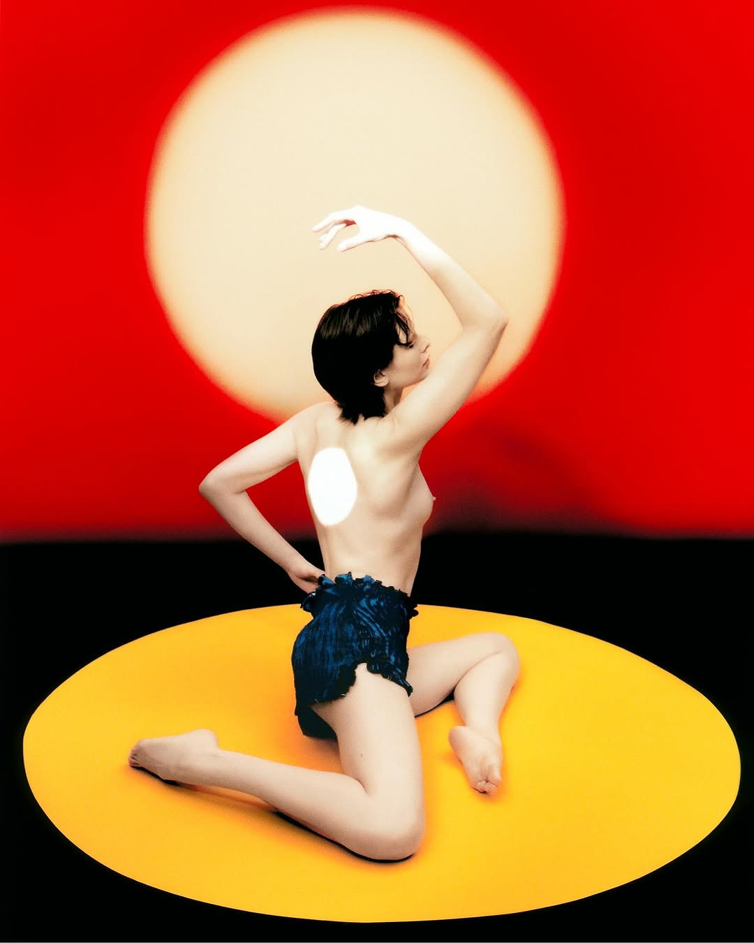 Backlit artistic nude portrait of a woman kneeling on a yellow platform against a red background with a large glowing circular light.